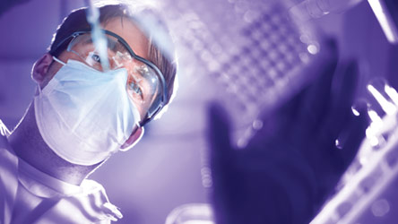 Лаборант с маска и защитни очила работи с лабораторно оборудване, фокусиран върху анализ и безопасност.