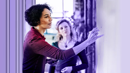 Жена обяснява идея на бяла дъска пред колега, демонстрирайки сътрудничество и креативно мислене в екипа.