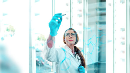 Учен с очила и ръкавици пише върху стъклена повърхност, демонстрирайки прецизност и научен подход.