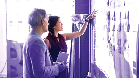 Двама колеги обсъждат информация на стена с диаграми, фокусирани върху стратегия и планиране.