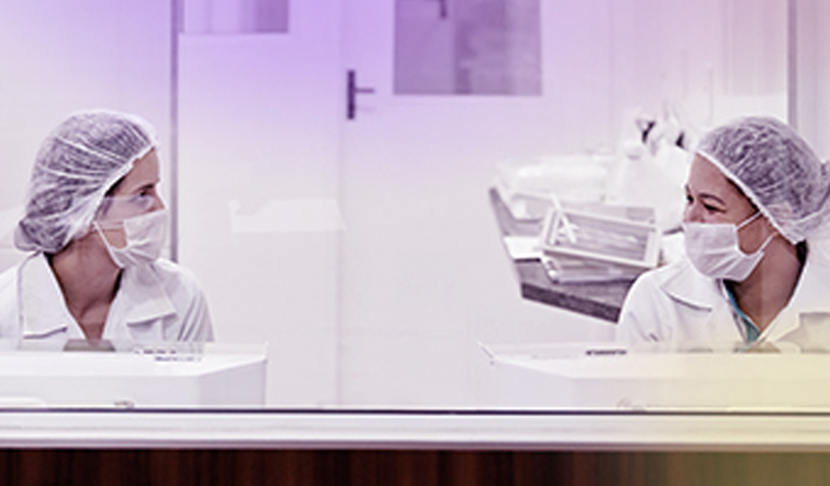 Две лаборантки с маски и защитни шапки разговарят в стерилна лабораторна среда, споделяйки опит и сътрудничество.