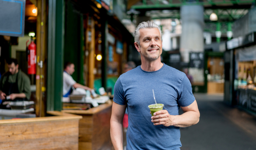 Усмихнат мъж с напитка в ръка се разхожда из пазара, наслаждавайки се на оживената атмосфера.
