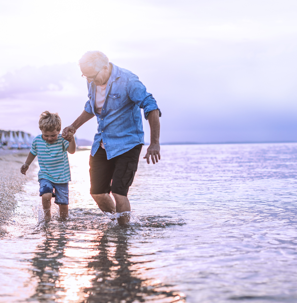 Възрастен мъж и малко момче се разхождат по плитката вода на плажа, държейки се за ръце в слънчев ден. 